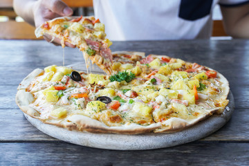 Man's hand holding a slice of Pizza with Mozzarella cheese, Ham, Tomatoes, salami, pepper, pepperoni. Pizza on wooden background.