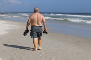 A man walking down the beach with his sandals in his hand and a camera in his hand looking at the incoming waves and a surfer approaching him