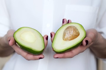 young woman in white shirt holding a two halves of fresh avocado in her hands