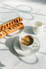 Coffee white cup, sweet rools on gray background in sunlight ray, selective focus. Breakfast concept, shadow, copy space