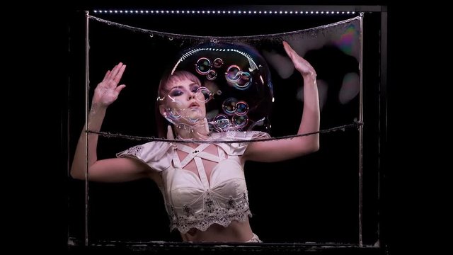 Pretty girl does tricks with soap bubbles. She blows a lot of small bubbles that are in big. With color highlights, on a vertical frame with LED lighting. Black background. Smiles