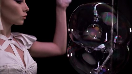 Close-up: Cute girl does tricks with soap bubbles. She blows lot of small bubbles into big bubbles, then removes them with her hand. With color highlights, on vertical frame with LED. Black background - Powered by Adobe