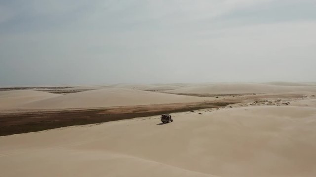 Aerial: A truck with kitesurfers traveling through the dunes of Lencois Maranhenses in Brazil, during dry season.