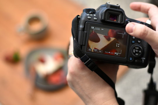 Hands of a photographer holding a camera, touching the screen with one finger taking photo of a cake on the plate and a cup of coffee in the blurred background. Closeup.