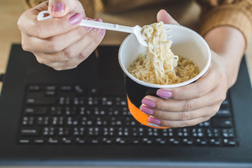 woman hand eating junk food noodles cup and working on laptop at office desk 