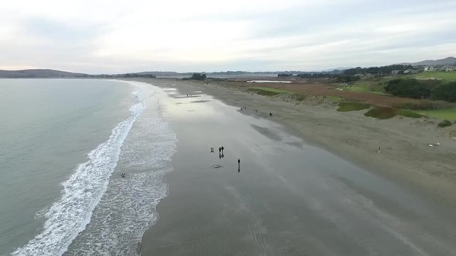 Moving shot of coastline with waves breaking along the shore, people walking in the distance, and a golf course sits adjacent