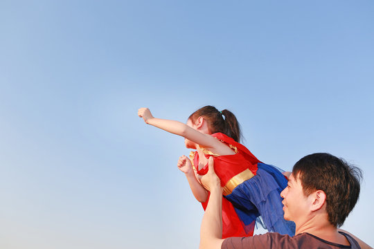 Smiling Asian father lifting up little cute child daughter in superhero dress flying having fun together on blue sky background. Kid girl enjoy playing with daddy. Super hero concept.