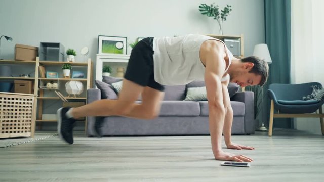 Side view of handsome young sportsman exercising at home doing cardio exercise in plank position using smartphone. Gadgets, millennials and house concept.