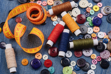 Fototapeta na wymiar A pile of multicolored sewing buttons, yellow measuring tape and thread spools on denim jeans background. Tailoring concept. Flat lay.