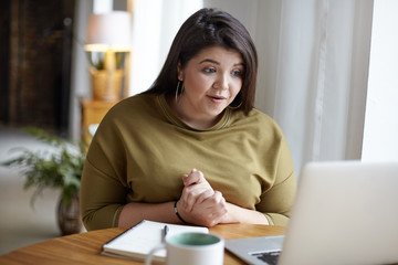 Adorable fashionable young plus size woman sitting at cozy cafeteria in front of open laptop, using...