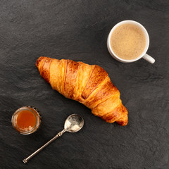 An overhead photo of a croissant with a cup of coffee and a jar of jam on a black background with copy space