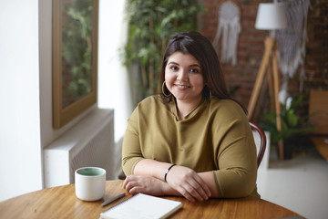 Horizontal shot of beautiful friendly young European woman with excess weight having coffee at cozy...