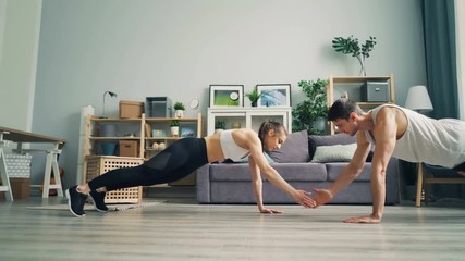 Young man and woman in sportswear are doing advanced pushups and clapping hands together in flat enjoying physical activity and pair work. Youth and fitness concept.