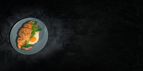Fresh french croissant sandwich with lettuce and fried egg on dark background. Delicious and healthy breakfast