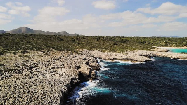a drone shot flying towards a rocky shore with waves crashing into the rocks and spraying into the air near cala varques in majorca