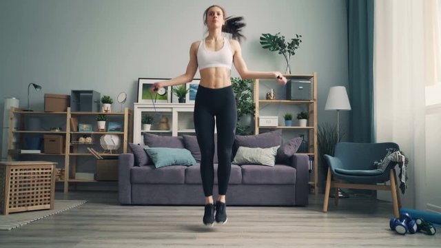 Slender girl in leggings and top is jumping rope at home concentrated on physical activity training legs losing weight. People, fitness and flat concept.