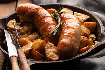 fried potatoes and grilled sausages in iron skillet with fork, knife near grey napkin and on wooden background