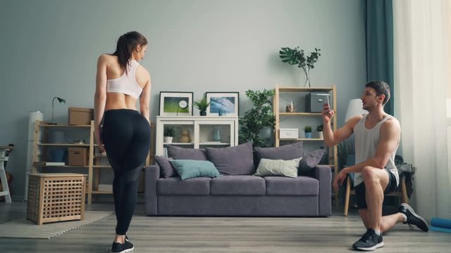 Young man is recording video of beautiful graceful sportswoman indoors at home using smartphone camera. Girl in sportswear is moving demonstrating perfect body.
