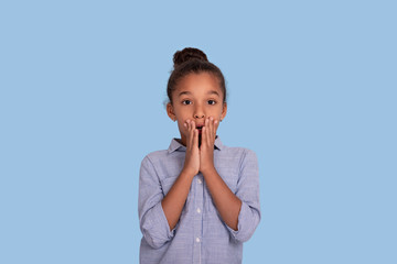 Close up emotional portrait of mulatta frizzy girl wearing blue shirt against blue background with copy space in studio. She covered her mouth with her hands in surprise.