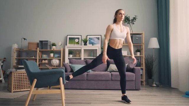 Beautiful young sportswoman is doing squats at home using armchair training body pumping muscles. Millennials, active people and interior concept.