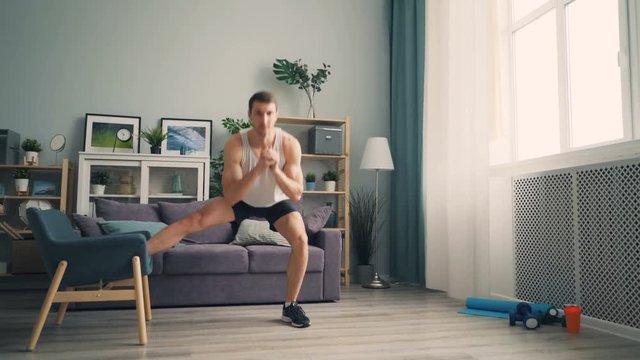 Young handsome man is squatting with armchair doing sports indoors at home working out leg muscles. Beautiful light apartment is visible in background.