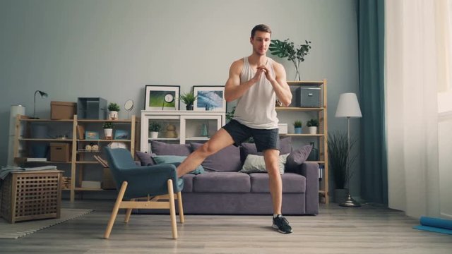 Handsome guy sportsman is doing squats with armchair in apartment enjoying sports wearing shorts, top and sneakers. Interior and active lifestyle concept.
