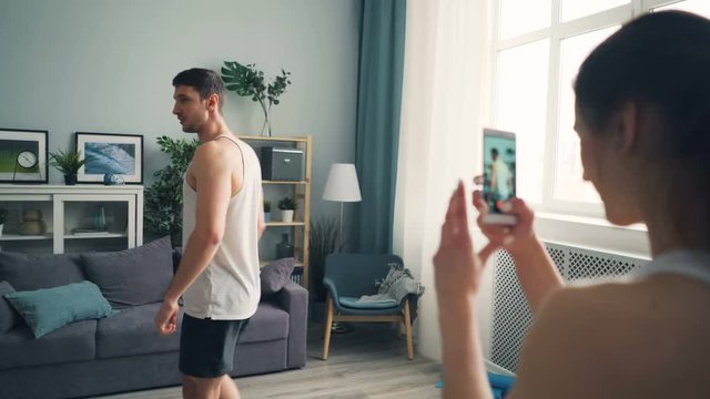 Handsome sportsman in shorts and top is posing for smartphone camera while girl is recording video touching screen. Modern technology and sports concept.
