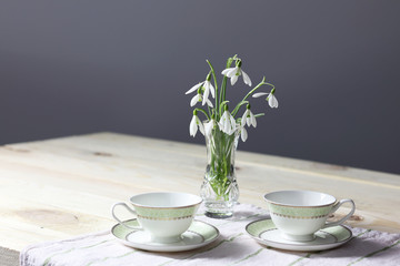 Snowdrops on the table. Spring flowers bouquet. Snowdrops background. Spring flowers on the wooden table. Holiday desk. Holiday flowers. Vase with spring flowers. Bowl with snowdrops. Two cups 