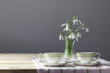 Snowdrops on the table. Spring flowers bouquet. Snowdrops background. Spring flowers on the wooden table. Holiday desk. Holiday flowers. Vase with spring flowers. Bowl with snowdrops. Two cups 