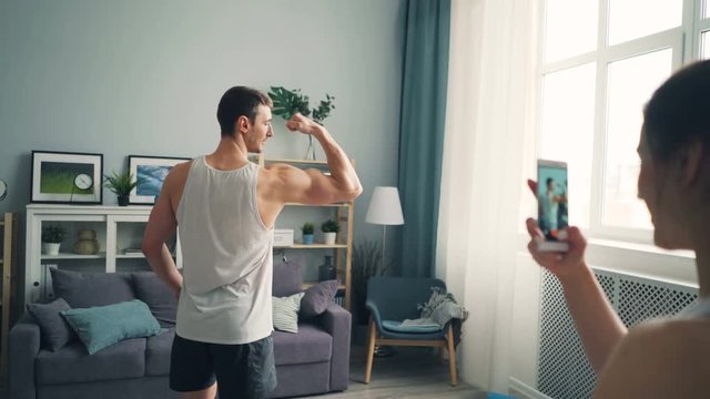 Young woman is recording video of handsome male bodybuilder using smartphone camera. Masculine guy in trendy sportswear is showing muscles posing and talking.