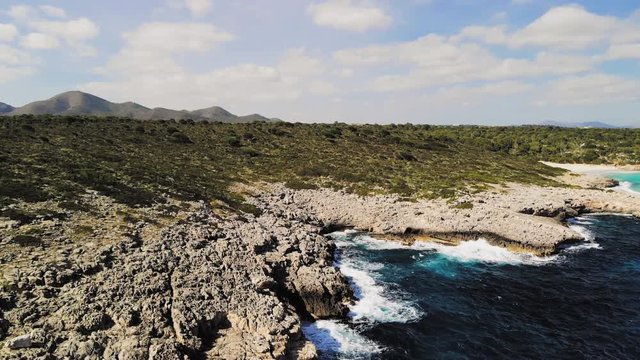 a drone shot flying over rocky shore with waves crashing into the rocks and spraying into the air near cala varques in majorca