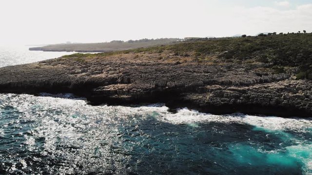 a drone shot of a rocky shore with waves crashing and slowly flying to pan up and out near cala varques in majorca