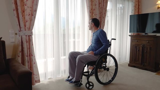 Depressed young man in wheelchair near the window at home or in nursing center. Slow motion 4k hand held movement