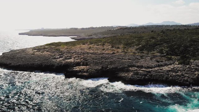 a forward flying drone shot of a large rocky landscape with waves crashing onto the rocks and creating sea spray near cala varques in majorca