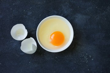 broken egg a whole bright yolk and white in a white plate and a white egg shell on a dark concrete background