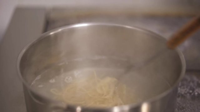 Handheld shot of putting and stirring handmade rolled spaghetti pasta in boiled water in a kitchen pot on the french cooking stove. Stirring spaghetti in a boiling water.