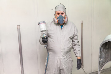 A male worker painting a car is standing in a spray booth in protective clothing with a spray gun in his hands pointing forward and looking into the camera