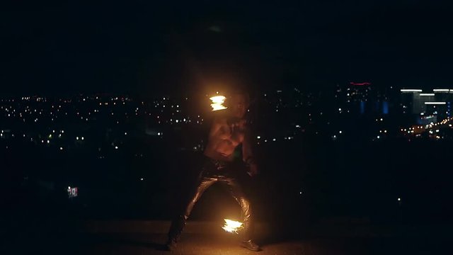 Young blond male naked torso spins two burning pois at night on black city background. Slow motion shot.