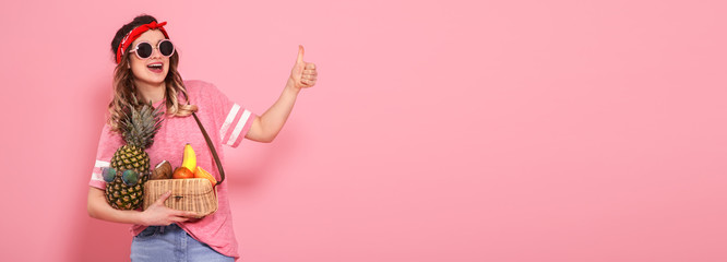 Beautiful young girl in pink t-shirt and glasses, holds a full straw bag of fruit on pink background. Shows finger class