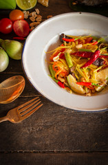 Papaya Salad (Som tum Thai) on white zine plate placed on the wood table there are eggplant, tomato, lemon, fork, spoon, bean, and chilli placed around.