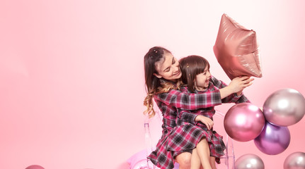 Obraz na płótnie Canvas Funny mom and child sitting on a transparent stylish chairs pink background. Little girl and mother having fun with balloons and confetti