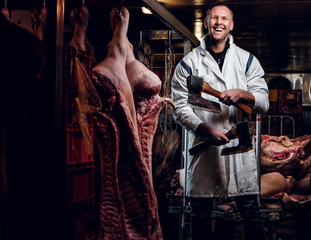 Cheerful butcher in workwear posing with two axes in a refrigerated warehouse in the midst of meat carcasses