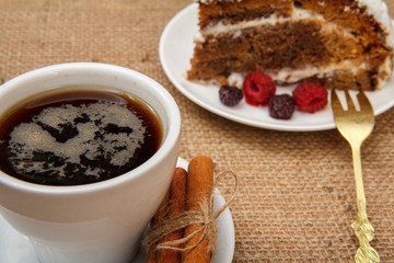 Cup of coffee, cinnamon, fork and slice of biscuit cake decorated with whipped cream and raspberries.
