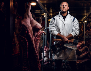 Fototapeta na wymiar The butcher in workwear posing with two axes in a refrigerated warehouse in the midst of meat carcasses