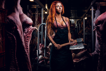 Redhead fitness woman in underwear and apron posing with ax in a refrigerated warehouse in the midst of meat carcasses