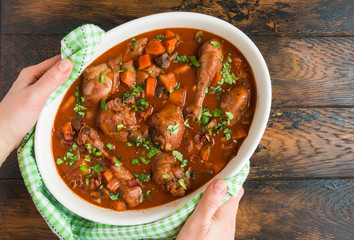 Coq au Vin, traditional French recipe of chicken braised in red wine with carrot and mushrooms. White casserole on wooden table, woman hands, top view