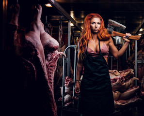 Redhead fitness woman in underwear and apron posing with ax in a refrigerated warehouse in the midst of meat carcasses