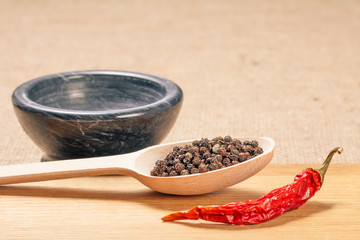 Wooden spoon with black peppercorns, dried red pepper on cutting board and stone mortar for grinding pepper