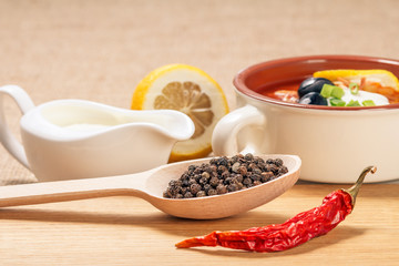 Wooden spoon with black peppercorn, dried red pepper on cutting board and ceramic soup bowl with saltwort, sauceboat and cut lemon.