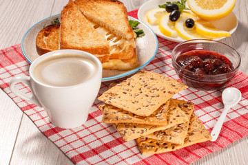 Toasted slices of bread with cheese and green parsley on white plate, cup of coffee, bowl with strawberry jam and spoon on napkin